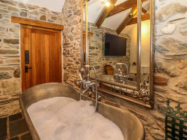 A bathroom with a stone bathtub filled with bubbles a wooden door and a large mirror at St Dogwells Barn near Wolfscastle