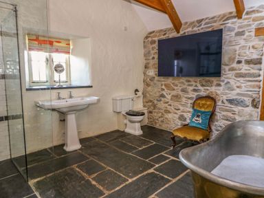 A bathroom with a stone wall, a pedestal sink, a toilet, a towel holder, a chair with a cushion, and a large metal bathtub at St Dogwells Barn near Wolfscastle
