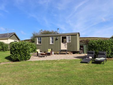 A small green corrugated cabin with outdoor seating and two lounge chairs on a grassy lawn at Shepherds Joy in Kennerleigh near Cheriton Fitzpaine