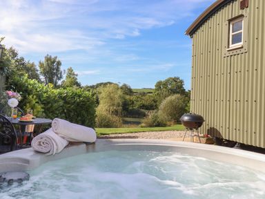 An outdoor hot tub with rolled towels on the edge a small round table with drinks and flowers a metal chair a green corrugated metal building a charcoal grill and trees in the background at Shepherds Joy Kennerleigh near Cheriton Fitzpaine