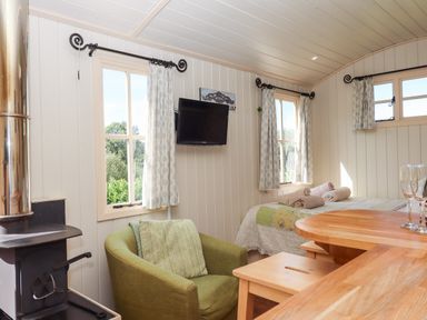 A small room with a wood stove a green armchair a bed with towels on it and a wall-mounted TV at Shepherds Joy in Kennerleigh near Cheriton Fitzpaine