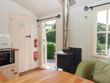 A small room with a wood stove a microwave a green chair and an open door showing greenery outside at Shepherds Joy in Kennerleigh near Cheriton Fitzpaine