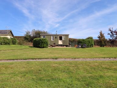 A small green cabin with a cream door and two chairs on a grassy lawn under a blue sky at Shepherds Joy in Kennerleigh near Cheriton Fitzpaine
