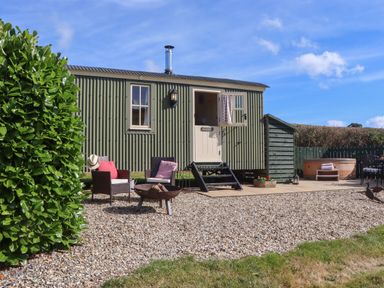 An outdoor seating area with chairs and a fire pit in front of a small green cabin with a door and windows at Shepherds Bliss Kennerleigh near Cheriton Fitzpaine