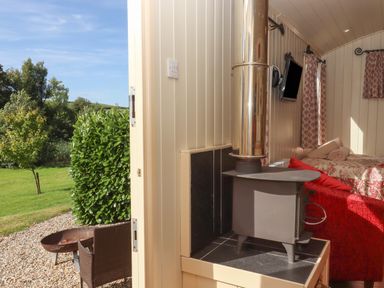 A small room with a wood stove a mounted TV a bed with patterned bedding and windows with curtains at Shepherds Bliss in Kennerleigh near Cheriton Fitzpaine