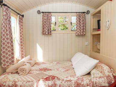A small bedroom with a bed covered in a patterned quilt and two white pillows with rolled towels on the bed and windows with curtains at Shepherds Bliss Kennerleigh near Cheriton Fitzpaine