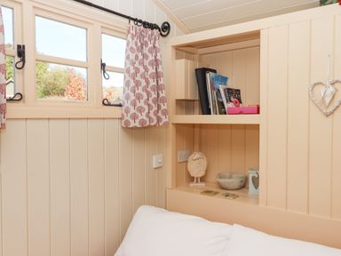 A corner of a room with a built-in shelf holding books and decorative items below a window with patterned curtains at Shepherds Bliss in Kennerleigh near Cheriton Fitzpaine