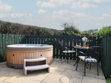 An outdoor patio with a hot tub and a table set with two chairs glasses orange juice pastries and flowers at Shepherds Bliss Kennerleigh near Cheriton Fitzpaine