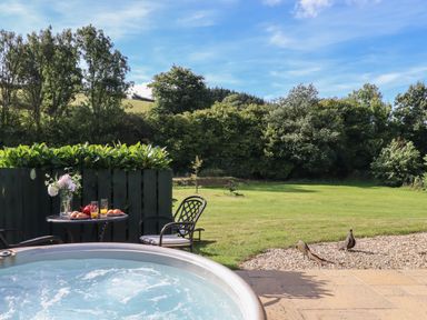 An outdoor patio with a hot tub a table with chairs and glasses on the table overlooking a grassy yard with trees at Shepherds Bliss Kennerleigh near Cheriton Fitzpaine
