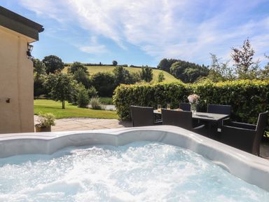 An outdoor hot tub and patio table with chairs and flowers with grassy hills and trees in the background at Binneford Barn Kennerleigh near Cheriton Fitzpaine