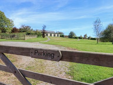 A wooden gate with a parking sign leading to a gravel driveway and a building in the distance with green lawns and trees at Binneford Barn Kennerleigh near Cheriton Fitzpaine