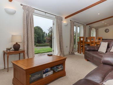 A living room with a wooden coffee table, a brown leather sofa, a dining table with chairs, curtains, and an open door showing an outdoor view at Binneford Barn Kennerleigh near Cheriton Fitzpaine