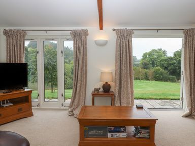 A living room with a wooden coffee table books a lamp a television on a wooden stand and large windows with curtains overlooking a garden at Binneford Barn in Kennerleigh near Cheriton Fitzpaine