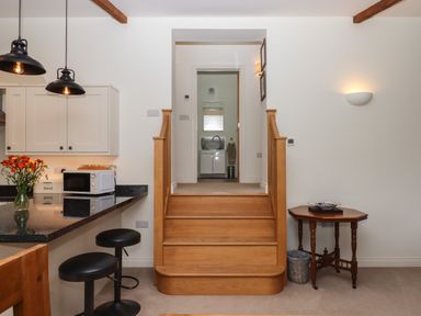 A kitchen area with black stools at a counter beside wooden steps leading to a room with a sink and mirror at Binneford Barn in Kennerleigh near Cheriton Fitzpaine