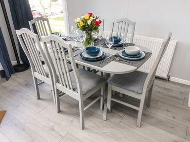 A dining area with a white oval table set with plates bowls glasses and cutlery six grey cushioned chairs and a vase of flowers on the table at Park View Lodge Arnside 7 South Lakeland Leisure Village near Carnforth