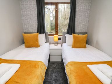 A bedroom with two single beds a nightstand with a lamp and a window with curtains at Park View Lodge Arnside 7 South Lakeland Leisure Village near Carnforth
