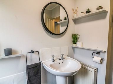 A bathroom with a round mirror a sink a towel rail with a towel two wall shelves with plants and a radiator at Park View Lodge Arnside 7 South Lakeland Leisure Village near Carnforth