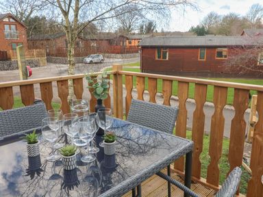 A balcony with a glass table set with wine glasses and small plants overlooking wooden cabins at Park View Lodge Arnside 7 South Lakeland Leisure Village near Carnforth