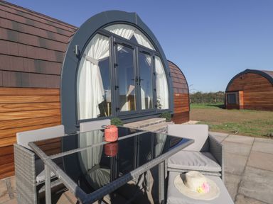 An outdoor patio with a glass table and cushioned chairs in front of a curved wooden cabin at Leadon View Oak in Upleadon near Newent