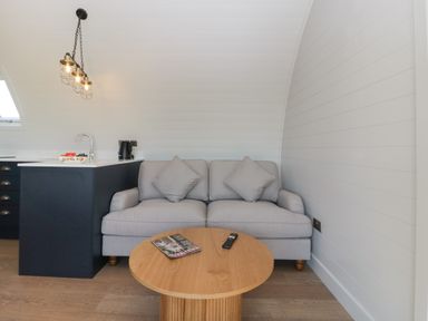 A small living area with a gray sofa two cushions a round wooden coffee table a magazine and a remote control on the table and a black kitchen island with a sink and a kettle at Leadon View Oak in Upleadon near Newent