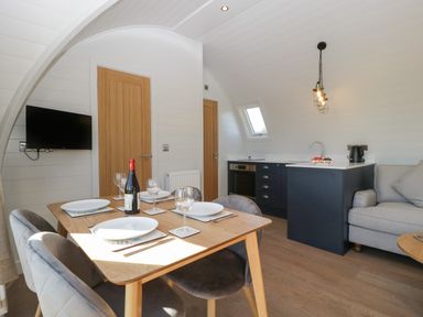 A dining table set for four with plates wine glasses and a bottle of wine near a kitchen with dark blue cabinets a sofa and a wall mounted television at Leadon View Oak in Upleadon near Newent