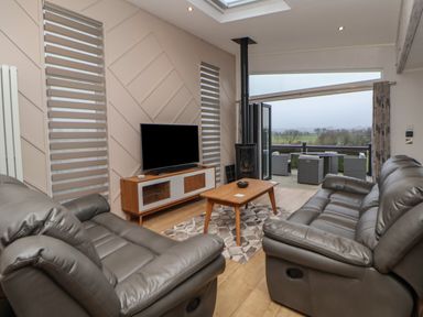A living area with two grey leather sofas a wooden coffee table a flat screen tv on a wooden stand and an open sliding door leading to an outdoor seating area with views of trees and fields at The Lodge @ Racecourse House in Babell near Holywell