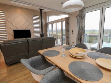 A dining area with a wooden table set with placemats and bowls next to a living room with a large leather sofa a wall-mounted TV and large glass doors at The Lodge @ Racecourse House in Babell near Holywell