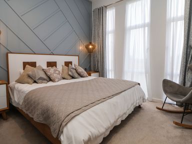 A bedroom with a bed covered with pillows and a blanket next to windows with curtains and a rocking chair at The Lodge @ Racecourse House in Babell near Holywell