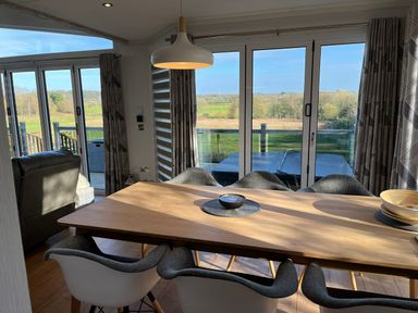 A dining area with a wooden table and six chairs next to large glass doors with a view of fields at The Lodge @ Racecourse House in Babell near Holywell