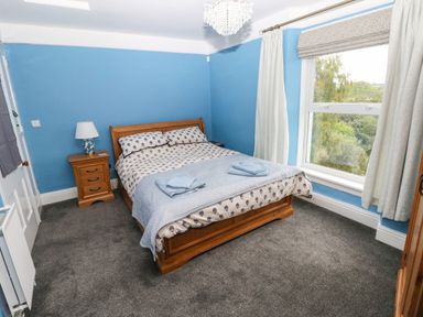 A bedroom with a wooden bed and nightstand a lamp on the nightstand blue walls and a window with curtains at Rushfield in St Dogmaels North Pembrokeshire