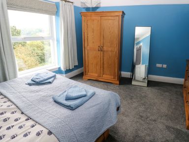 A bedroom with a bed covered in blue bedding a wooden wardrobe a standing mirror and a window at Rushfield in St Dogmaels North Pembrokeshire