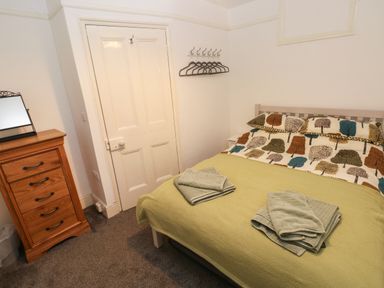 A bedroom with a bed covered with a tree patterned duvet and green blanket with towels a wooden chest of drawers with a mirror on top and a coat rack with hangers on the wall at Rushfield in St Dogmaels North Pembrokeshire