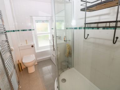 A bathroom with a toilet, a shower enclosure with glass doors, a small sink with a cabinet, a window with blinds, a towel rack, and a wall-mounted shelf at Rushfield in St Dogmaels North Pembrokeshire