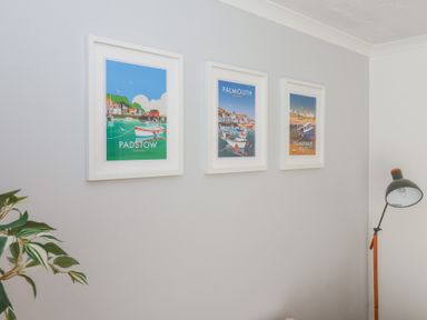 Three framed pictures on a wall featuring Padstow Falmouth and Summerleaze Beach next to a floor lamp and plant at Beachcomber Maer near Bude