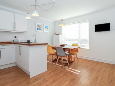 A kitchen and dining area with a wooden table and four chairs and a wall mounted television at Beachcomber in Maer near Bude
