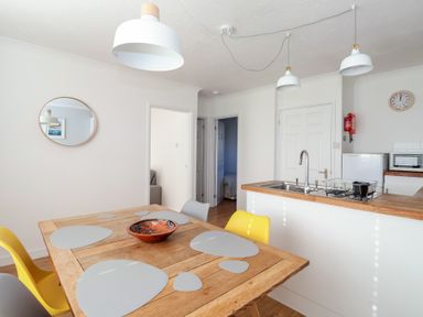 A dining area with a wooden table and placemats near a kitchen with a sink and hanging lights at Beachcomber Maer near Bude