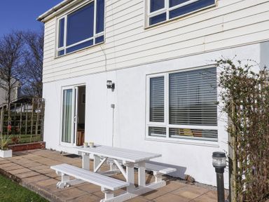 A white outdoor picnic table and benches on a patio outside a white house with windows and a sliding door at Beachcomber Maer near Bude