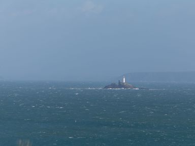 A lighthouse on a rocky islet in the ocean with distant cliffs on the horizon at Chalet 3 Wipeout in Hayle