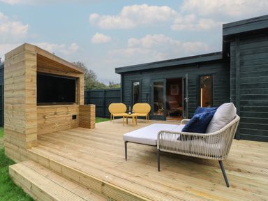 An outdoor wooden deck with a television, two yellow chairs and table with drinks and a lounge chair with cushions at Badger's Burrow in South Luffenham near Edith Weston