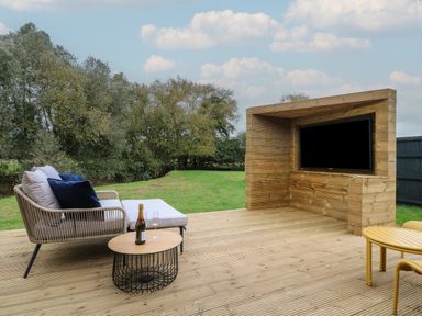 An outdoor wooden deck with a sofa, round table with a bottle and glasses, and a built-in wooden TV unit at Badger's Burrow in South Luffenham near Edith Weston