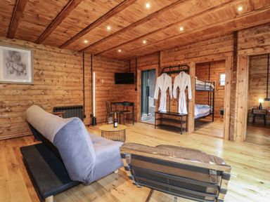 A living area with wooden walls and ceiling containing a couch, chair, round table with wine, TV on wall, two white robes hanging and visible bedrooms at Badger's Burrow South Luffenham near Edith Weston