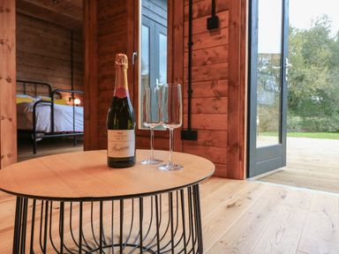 A wooden room with a small round table holding a bottle of prosecco and two glasses near an open door and a bedroom in the background at Badger's Burrow South Luffenham near Edith Weston