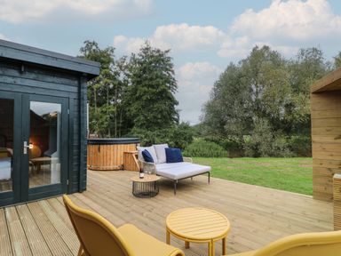 An outdoor wooden deck with lounge chairs small tables a sofa with cushions and a hot tub near trees at Badger's Burrow in South Luffenham near Edith Weston