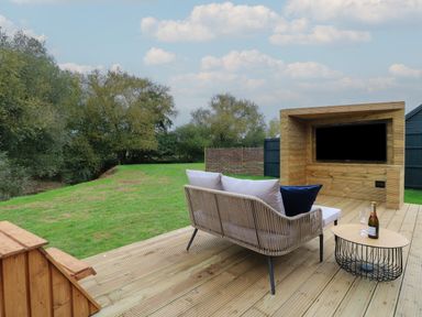 An outdoor wooden deck with a sofa facing a wooden entertainment unit with a TV and a round table with a bottle and glass at Badger's Burrow South Luffenham near Edith Weston
