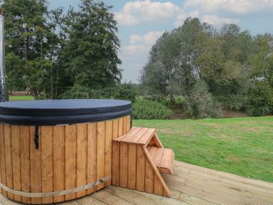 An outdoor wooden hot tub with steps on a wooden deck surrounded by grass and trees at Badger's Burrow in South Luffenham near Edith Weston
