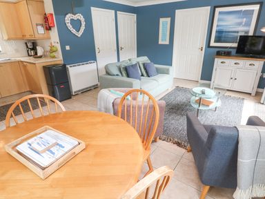 A living room with blue walls a light green sofa a dark blue armchair a glass coffee table a wooden dining table and kitchen cabinets at Chy Lowena Goldenbank near Budock Water