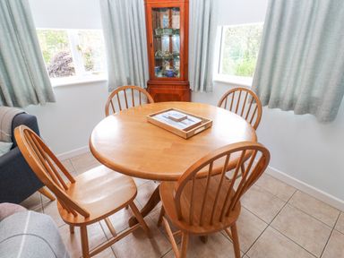 A dining area with a round wooden table and four wooden chairs with curtains on windows at Chy Lowena Goldenbank near Budock Water