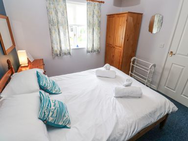 A bedroom with a double bed two pillows a wooden wardrobe a window with curtains and rolled towels on the bed at Chy Lowena in Goldenbank near Budock Water