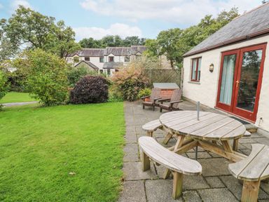 A patio with a round wooden picnic table and chairs next to a house with red doors and windows and a grassy garden with bushes at Chy Lowena Goldenbank near Budock Water