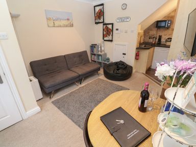 A living room with a gray sofa a round wooden table a black bean bag and a kitchenette at 5 Lyonesse Apartments in St Ives